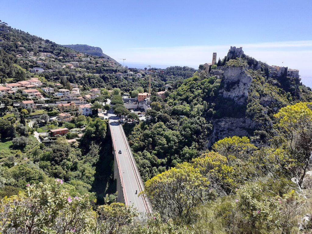 A car travelling the Moyenne Corniche D6007 with the medieval village of Eze perched on the limestone ridge above and the Mediterranean coast stretching toward Monaco in the distance.